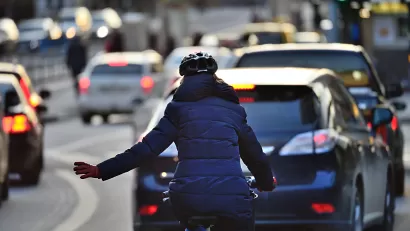 Rear view of woman in blue coat riding bike on busy road signals with left hand to merge into lane of traffic.